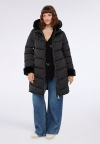 Schwarze Steppjacke mit Kapuze, ausgestattet mit Kunstpelzärmeln und goldfarbenem Reißverschluss. Tragen über einem schwarzen Oberteil, kombiniert mit weit geschnittenen Jeans und hellen Schuhen.