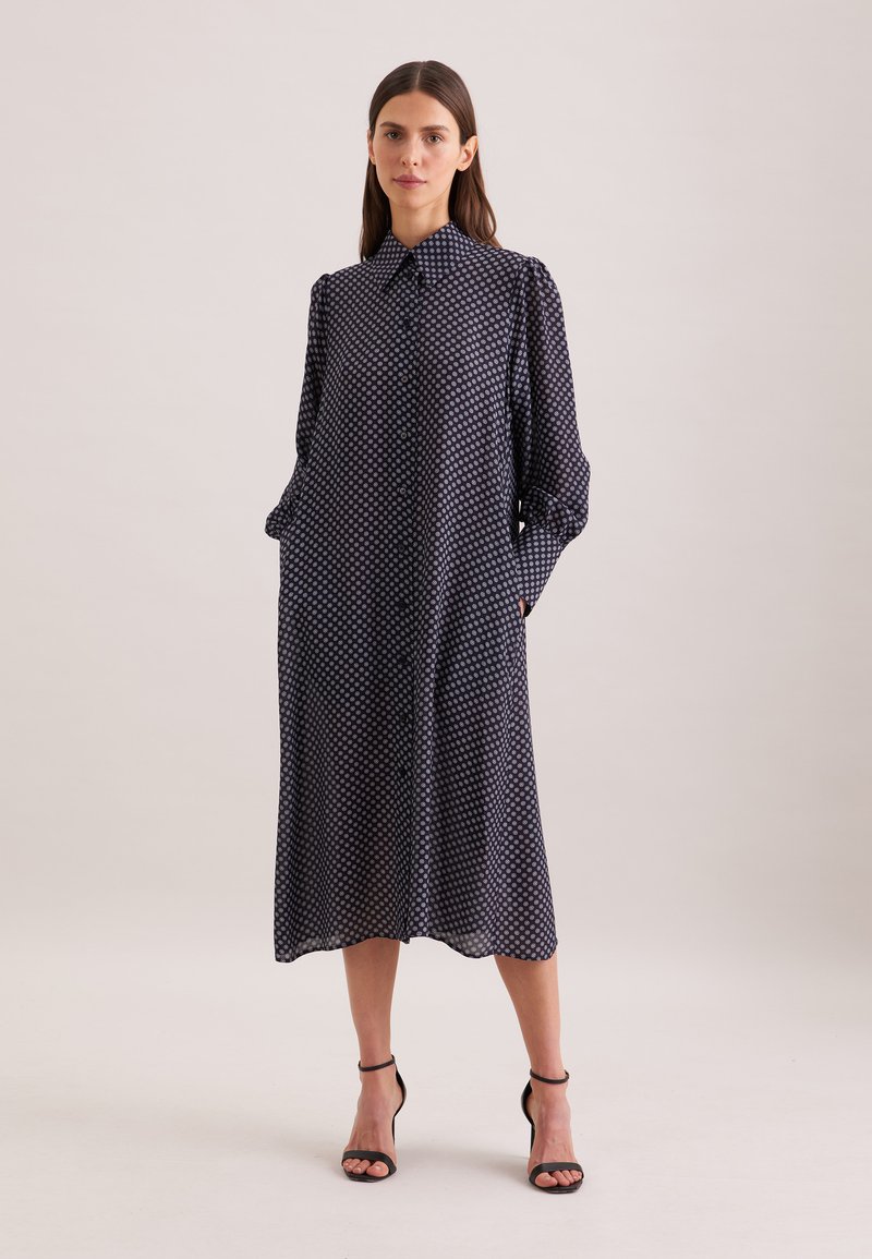 Une femme porte une robe midi bleu marine à pois blancs, manches longues, boutonnée sur le devant, avec des talons noirs à brides autour de la cheville.