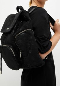 Zaino nero con motivo logo in rilievo, materiale in tessuto, tasca frontale con zip, chiusura a cordoncino e spallacci regolabili.