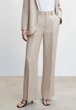 Pantalons en lin beige clair avec un design à jambes larges, dotés de plis à l'avant, d'une taille haute et d'une texture subtile. Associés à des sandales noires à lanières.