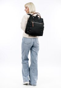 Woman with short blonde hair wearing a beige sweater, blue jeans, white sneakers, and carrying a black backpack with brown zipper pull.