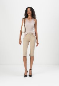 Top de malha bege canelada com rendas, combinado com calças capri bege claro e sapatos de salto agudo pretos; a modelo segura uma mala de ombro creme.