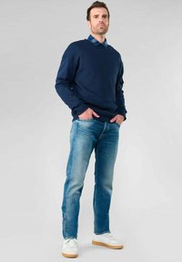 Maglione blu navy sovrapposto a una camicia a quadri, abbinato a jeans azzurri chiari e sneakers bianche con suola beige. Design semplice e casual.