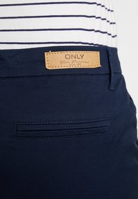 Tmavě modré denimové kalhoty s koženým štítkem značky "ONLY" na pasu, s detailem zadní kapsy a hladkou texturou.