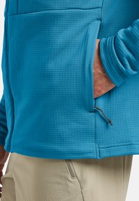 Maglia blu a maniche lunghe con texture, dotata di tasca laterale con chiusura a coulisse e dettagli ai polsini, abbinata a pantaloni beige.