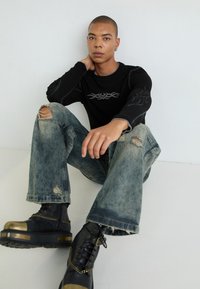 Schwarzes, geripptes Langarmshirt mit metallischem Grafikdesign; hellblaue, ausgebeulte Jeans mit weiter Beinform; schwarze Stiefel mit goldenen Akzenten und Kettendetails.