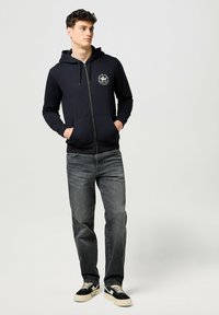Navy zip-up hoodie with a front kangaroo pocket and logo detail on the chest, paired with gray jeans and black shoes.