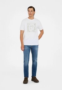 Weißes T-Shirt mit kurzen Ärmeln und grafischem Design, kombiniert mit blauer Jeans und braunen Schuhen, vor neutralem Hintergrund stehend.