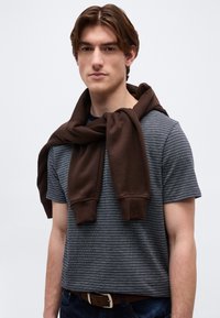 Νέος άνδρας με καστανά μαλλιά, που φοράει γκρι ριγέ μπλουζάκι και καφέ φούτερ δεμένο γύρω από τους ώμους του, πάνω σε απλό φόντο.