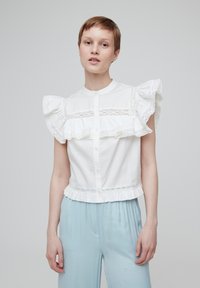 Weiße Bluse mit kurzen Ärmeln, verziert mit gerafftem Spitzenbesatz, Knopfleiste vorne und fransigem Saum. Kombiniert mit hellblauen Hosen.