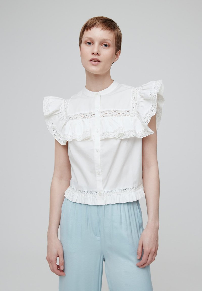Weiße Bluse mit kurzen Ärmeln, verziert mit gerafftem Spitzenbesatz, Knopfleiste vorne und fransigem Saum. Kombiniert mit hellblauen Hosen.
