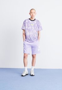 Gaiši purpura krāsu sānu svītru T-krekls ar melnu apkakli un dzeltenu akcentu. Apvienots ar atbilstošām purpura šortiem, kuros ir balts Nike logo. Baltas sporta kurpes.