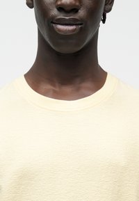 Gros plan sur le bas du visage et le cou d'une personne portant un pull texturé col rond de couleur crème, sur fond uni.