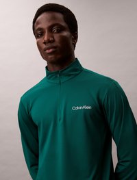 Zelená zip-up dlhá rukávová blúzka s text�úrovanou tkaninou. Obsahuje vysoký golier a biely logo "Calvin Klein" na ľavej hrudi.