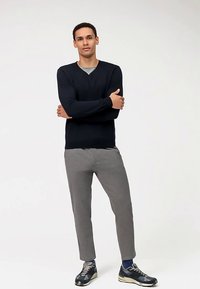 Marineblauwe v-hals trui, grijze slim-fit broek en donkere sneakers. Model staat met gekruiste armen tegen een effen achtergrond.