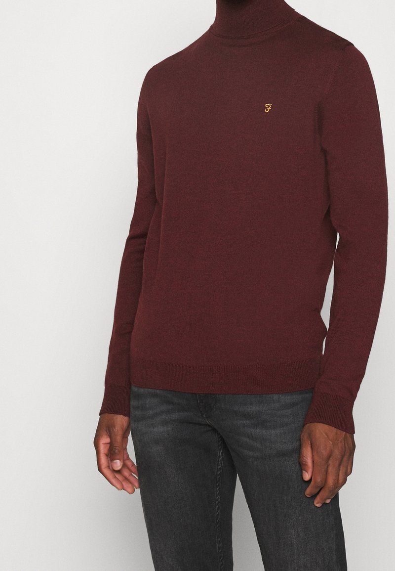 Burgunderroter Rollkragenpullover aus weichem Strickstoff, mit gerippten Bündchen und Saum, versehen mit einem kleinen goldenen Logo auf der Brust.