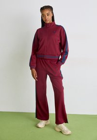 Bordeaux atletisk tracksuit med lynlåsjakke og elastisk talje. Har blå sidelommer og logodetaljer. Båret med beige sneakers.