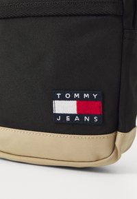 Fekete anyagú hátizsák, amelynek alja bézs színű. Tartalmaz egy téglalap alakú hímezett foltot "TOMMY JEANS" felirattal és színes téglalap mintával.