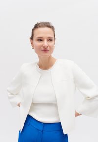 Witte jas met driekwartmouwen over een geribbeld wit topje; gecombineerd met felblauwe high-waisted broek; minimalistisch ontwerp.