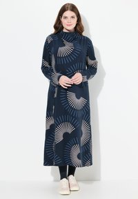 Långärmad marinblå klänning med ett fläktmönster i beige och ljusblått. Slät textur, hög halsringning, kombineras med svarta leggings och ljusa sneakers.