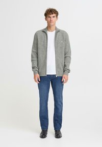 Pelēks adīts džemperis ar rāvējslēdzēju un ribotu tekstūru, uzvilkts virs baltas krekla. Saskaņots ar ziliem džinsiem un melnām kurpēm. Modelis stāv uz balta fona.