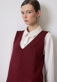 Burgundy stickad väst med V-ringning, bärs över en vit skjorta med krage. Har en slät textur och enkla sömdetaljer.