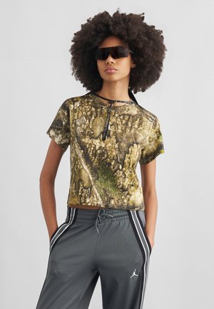 Nuori henkilö, jolla on suuret kiharat hiukset, käyttää tummia naamioivia aurinkolaseja, maastokuvioista lyhytpaitaista t-paitaa ja harmaita urheiluhousuja valkoisin raidoin.