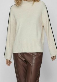 Crème ribbeltrui met een hoge hals en zwarte zijstrepen. Gecombineerd met bruine leren broeken, die een relaxte pasvorm en textuur hebben.