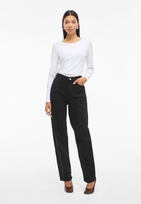 Crne hlače visokog struka s ravnim nogavicama, izrađene od teksturirane tkanine. Uparene s jednostavnim dugim bijelim topom i smeškim potpeticama.