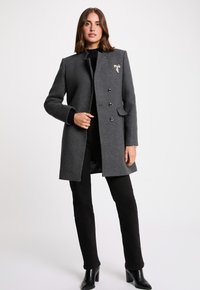 Grau wollener Mantel mit einem kragenlosen Design, drei Knöpfen und Seitentaschen. Akzentuiert mit einer dekorativen Schleife auf der Brust. Schwarze Hose und High Heels.