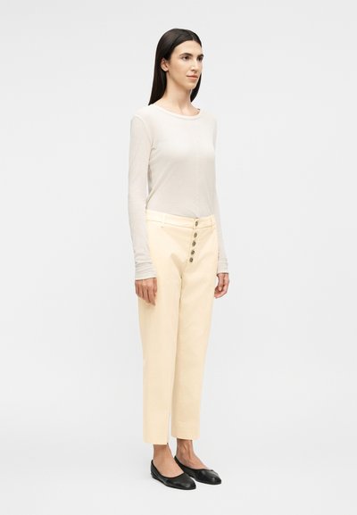 Γυναίκα που στέκεται φορώντας ένα διάφανο λευκό κορμάκι με μακριά μανίκια, κρεμ ψηλόμεσα παντελόνια με κουμπιά και μαύρες μπαλαρίνες σε απλό φόντο.