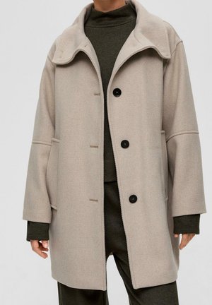 Beige wollen jas met brede kraag en zwarte knopen, gedragen over een donkergroene coltrui en bijpassende broek, handen ontspannen langs het lichaam.