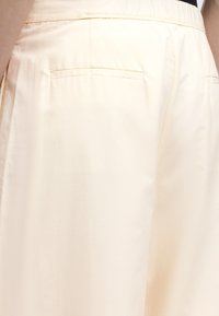 Κοντινό πλάνο σε κρεμ παντελόνια με πίσω τσεπάκια και ραμμένη ζώνη, φορεμένα με μαύρο τοπ.