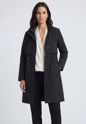 Nainen, jolla on pitkät tummat hiukset, pukeutuneena mustaan pitkä takkiin valkoisen paidan ja mustien housujen päällä, seisoo yhdellä kädellä taskussa.