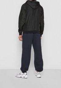 Zwarte lichte jas met een capuchon, elastische manchetten, gecombineerd met marineblauwe joggingbroek en witte sneakers, die een ontspannen pasvorm en eenvoudig ontwerp laat zien.