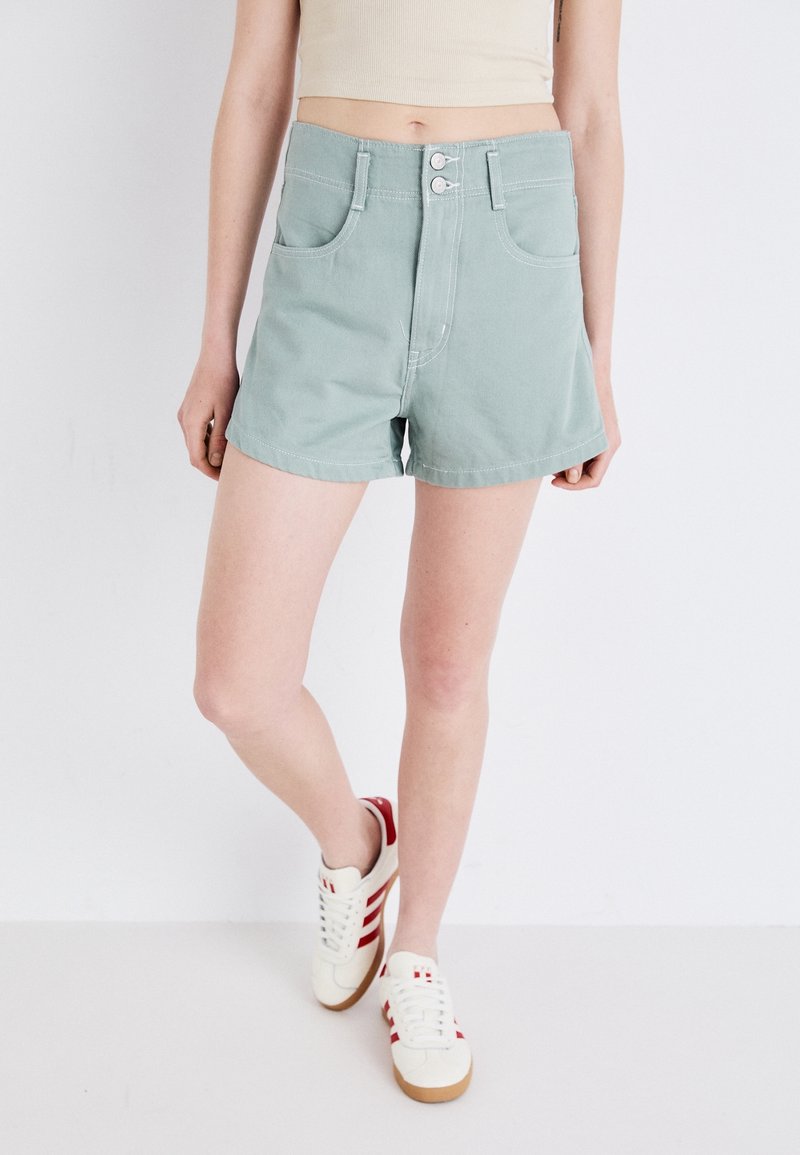 Femme portant un short taille haute vert clair avec des boutons à l'avant et des baskets blanches avec des bandes rouges, debout sur un fond blanc uni.