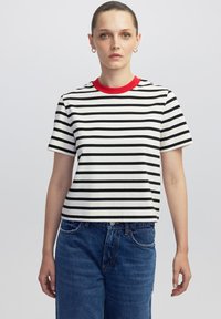 Mujer vistiendo una camiseta de rayas blancas y negras de manga corta con un collar rojo y vaqueros azules, de pie contra un fondo simple.