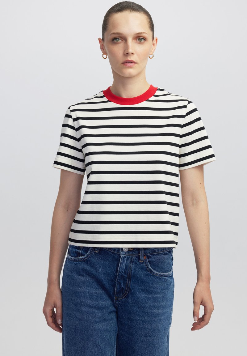 Mujer vistiendo una camiseta de rayas blancas y negras de manga corta con un collar rojo y vaqueros azules, de pie contra un fondo simple.