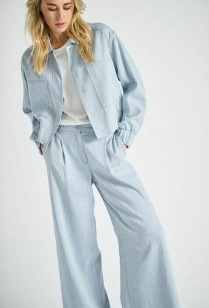 Vrouw die lichtblauwe broek met wijde pijpen en bijpassend jasje met knopen draagt over een wit T-shirt, handen in de zakken, staand tegen een effen achtergrond.