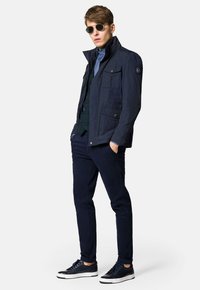 Granatowa kurtka z wysokim kołnierzem, wieloma kieszeniami i gładką teksturą, połączona z granatowymi spodniami i ciemnymi sneakersami z szarymi akcentami.