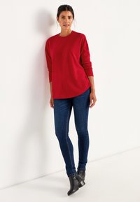 Rode geribde trui met lange mouwen, gebogen zoom, gedragen met donkerblauwe skinny jeans en zwarte enkellaarzen, staand tegen een witte achtergrond.