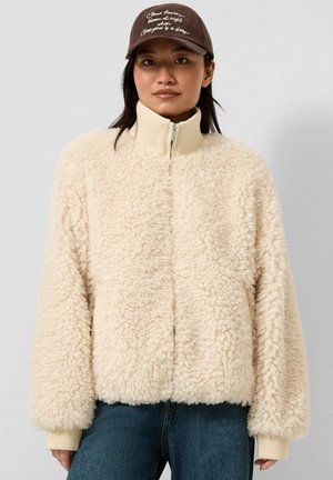 Frau trägt eine cremefarbene, flauschige Jacke mit Reißverschluss, blaue Jeans und eine braune Mütze mit weiß besticktem Text.