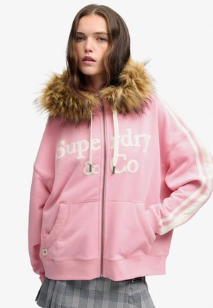 Jonge vrouw draagt een roze zip-hoodie met bontkraag rond de capuchon en een grijze geruite rok, staand met één hand in de zak.