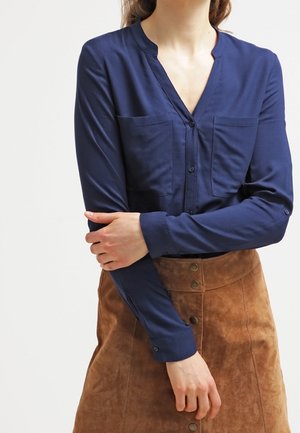 Femme portant un chemisier bleu marine à boutons avec deux poches poitrine et une jupe marron en daim boutonnée devant, les bras croisés.