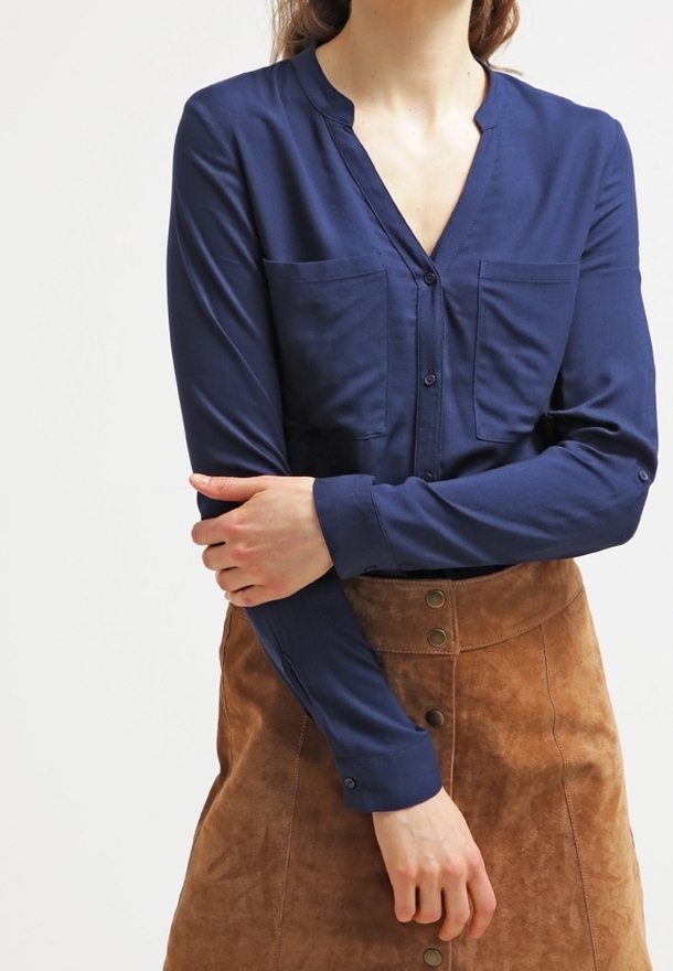 Femme portant un chemisier bleu marine à boutons avec deux poches poitrine et une jupe marron en daim boutonnée devant, les bras croisés.