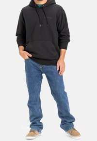 Uomo che indossa un felpa con cappuccio nera, jeans blu e sneakers color beige, in piedi con una mano in tasca contro uno sfondo bianco.