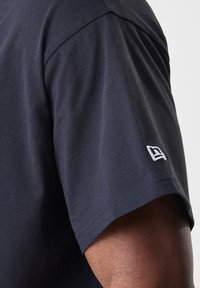 Námornícky modré bavlnené tričko s krátkymi rukávmi, s bielou vyšitou nášivkou na rukáve a hladkou, rovnomernou textúrou.