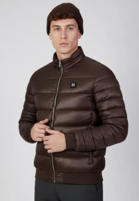 Chaqueta acolchada marrón con un cuello alto, diseño acolchado horizontal, cremallera frontal y un logo de marca negro en el pecho.