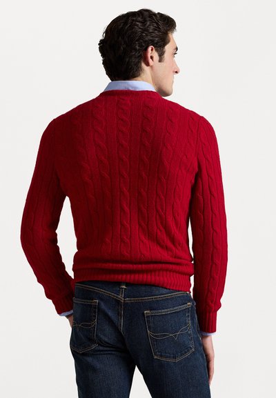 Suéter rojo de punto con trenzas, con puños y dobladillo acanalados, llevado encima de una camisa de cuello azul claro, combinado con un pantalón de mezclilla oscuro.
