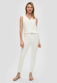 Witte mouwloze top met bloemenborduring, V-hals, gecombineerd met slim-fit witte broeken en platte sandalen met enkelbandjes.